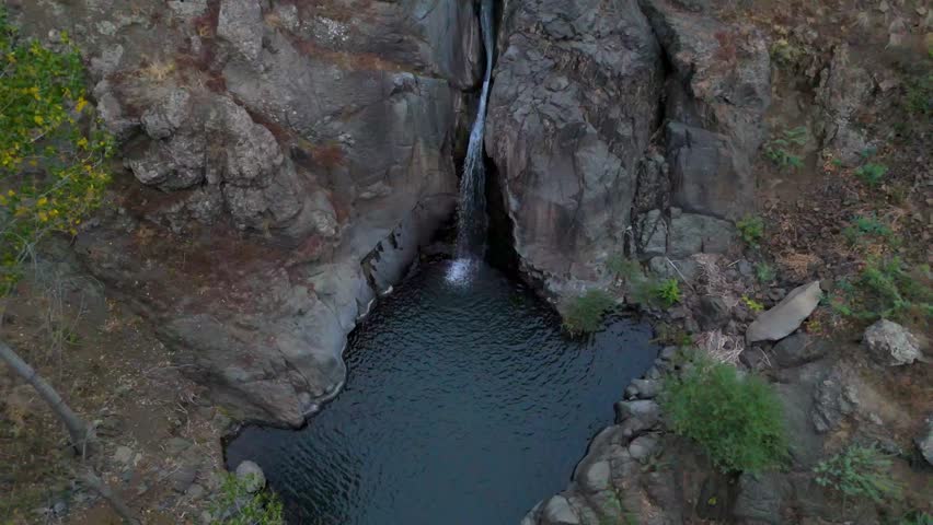 Aerial view of a small waterfall cascading into a natural rock pool surrounded by rugged cliffs and sparse vegetation. Herher Waterfall