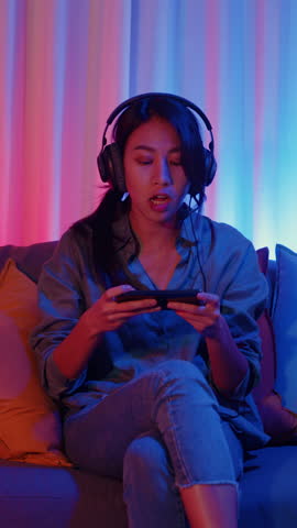 Vertical Screen : Happy asia girl gamer wear headphone competition video game online with smartphone excited talk with friend sit on couch in colorful neon lights living room at home, Home activity.