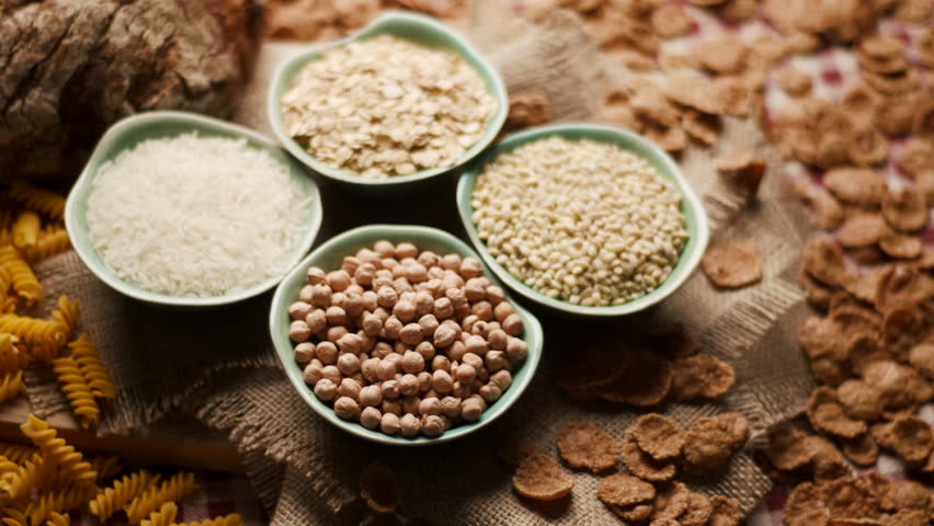 Close-up of carbohydrate-rich foods on a table, including bread, rice, wholegrain flakes, oats, wheat and corn pasta. Carbs Food.