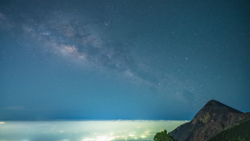 Time lapse of the Milky Way stretching above the active Fuego Volcano in Guatemala, with glowing city lights below and a vivid, starry night sky. - Powered by Shutterstock - Get 15% off with code: PIKWIZARD15
