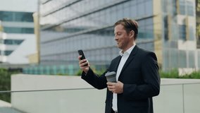 Portrait of businessman checks market trends via smartphone and smiles by skyscrapers. Mature entrepreneur sends message by cellphone holding coffee to go in modern downtown block - Powered by Shutterstock - Get 15% off with code: PIKWIZARD15