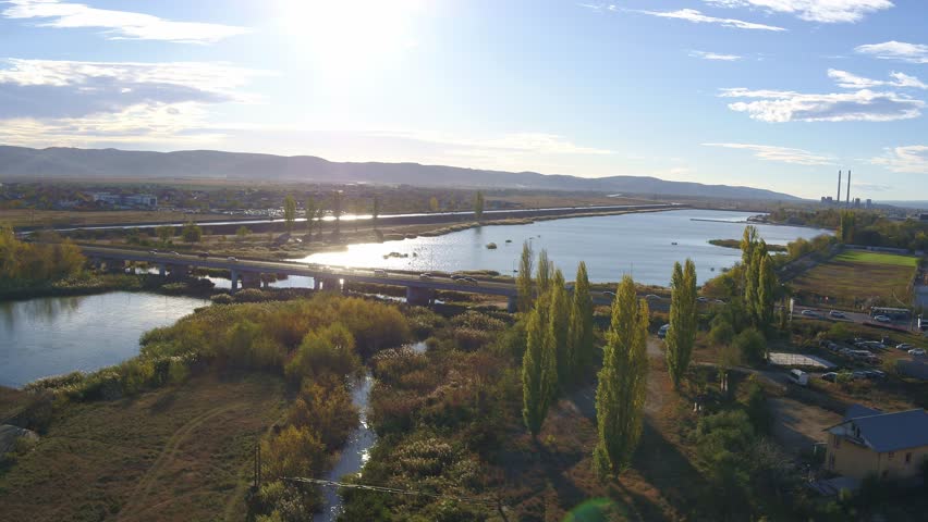 A slow-panning aerial shot captures a scenic river landscape in Bacau during golden hour. The setting sun reflects on the water as traffic crosses a bridge, with a power plant behind.