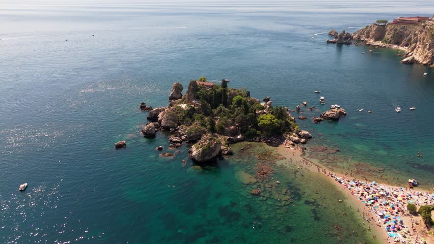 Drone slowly pans right to left around Isola Bella, showcasing the island, coastline, and sandy beach in a serene aerial view.