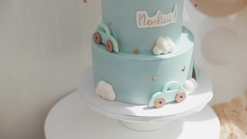 A two-tiered, light blue birthday cake with toy cars, clouds, a teddy bear, and 'Noeliui ' written on it, with nearby balloons