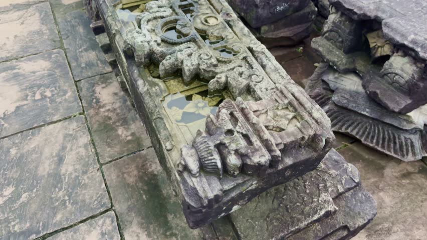 A slow push-in reveals a weathered carved stone fragment, its surface etched with ornate floral and scroll motifs, rainwater resting in its grooves, echoing ancient artistry and timeless devotion.