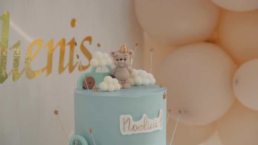 A two-tiered, light blue birthday cake with toy clouds, a teddy bear, and 'Noeliui ' written on it, with nearby balloons