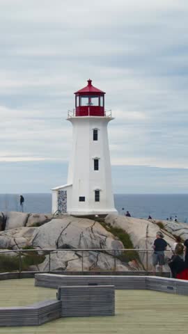 Vertical Video, Large Crowd Of Tourists Moves Quickly Along The Rocky Shore By The Famous Lighthouse In Peggys Cove, Nova Scotia. Time-Lapse Captures Mass Movement Against Stunning Ocean Backdrop.