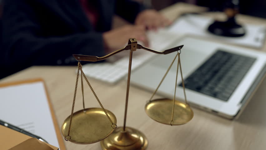 Golden justice scales in focus with blurred lawyer typing on keyboard, laptop, and legal tools in background. Concept of fairness, law, and digital legal services.