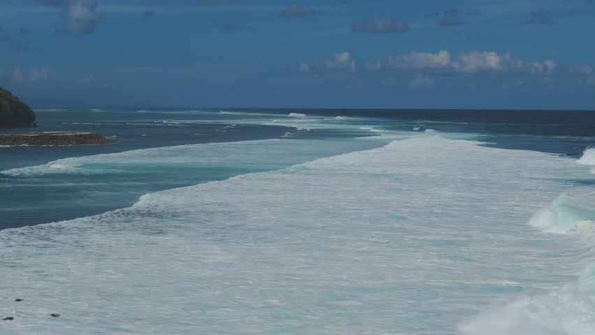 Drone lateral move follows whitewater bands over the reef near Melasti Beach, Bali, with deep blue sea and open horizon under fair weather. Daytime clarity highlights wave rhythm and coastal power