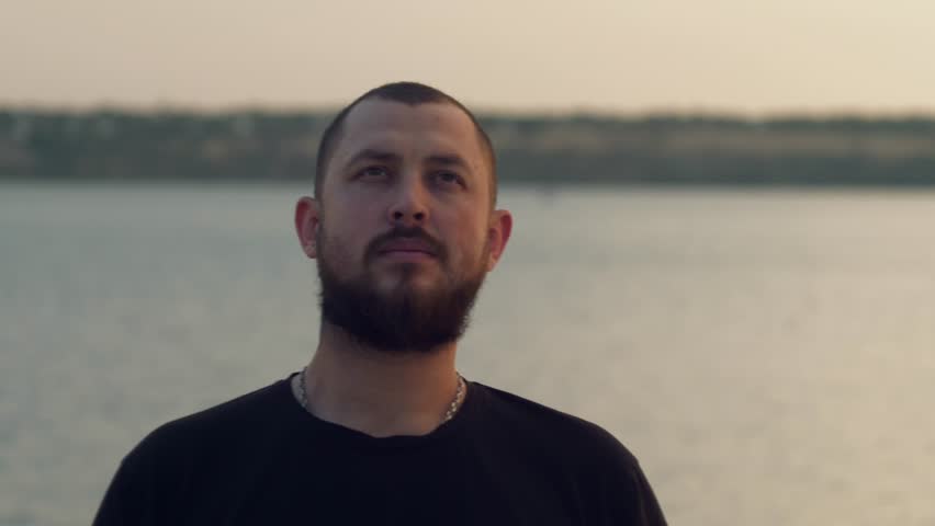 Cinematic parallax shot of a young bearded man standing by the seaside during sunset. The camera moves around him as he looks into the distance, creating depth and emotional atmosphere.