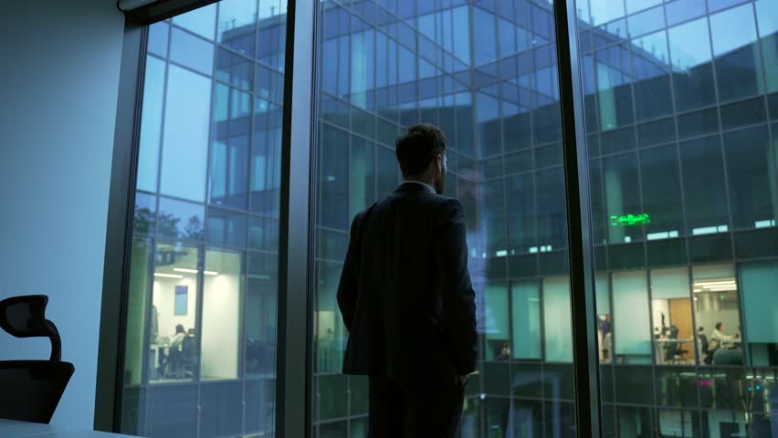Businessman looking out the window of a modern business office. Dark silhouette. Rear view. Successful thoughtful manager in a formal suit standing in the workspace and contemplating the cityscape