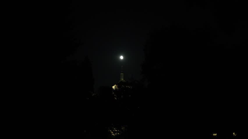 Bright full moon shines above dark silhouette of trees with illuminated church tower visible below creating peaceful night scene filled with mystery and quiet spiritual atmosphere