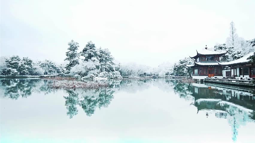 Snow falls gently on a serene winter landscape. A traditional pagoda and snow-covered trees are perfectly reflected in the still water of a calm lake.