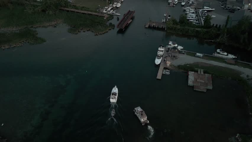 A drone view of two boats cruising along a dark canal near a rusted bridge and docked vessels at a marina in Orillia, Ontario on a summer evening