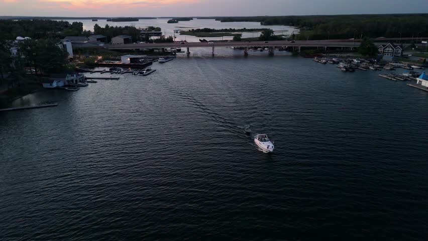 A drone view passing a white boat on dark water at sunset, revealing a narrow harbor with docks, buildings, and a traffic bridge in Orillia, Ontario