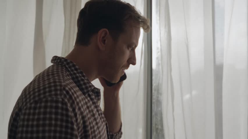 Man making phone call while standing by window, sunlight shining through. Guy talks on his smartphone, frustrated and upset expression, difficult or emotional conversation, stress, conflict, bad news.