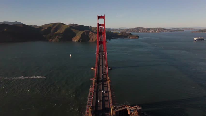 Drone flies low over the Golden Gate Bridge in San Francisco, with cars below and a sailboat, motorboat, and large ship visible in the bay beyond.