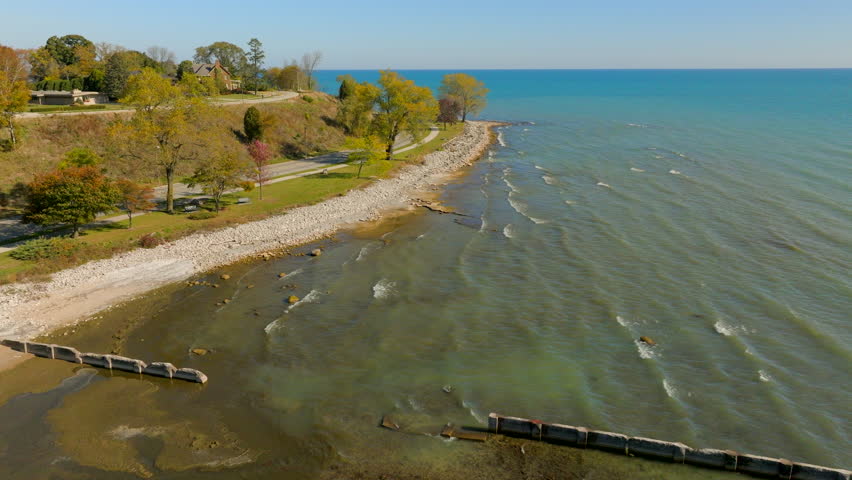 Drone aerial flying above Lake Michigan shoreline in Sheboygan, Wisconsin with cars along the road, colorful autumn trees, and neighborhoods on a bright, pretty day.