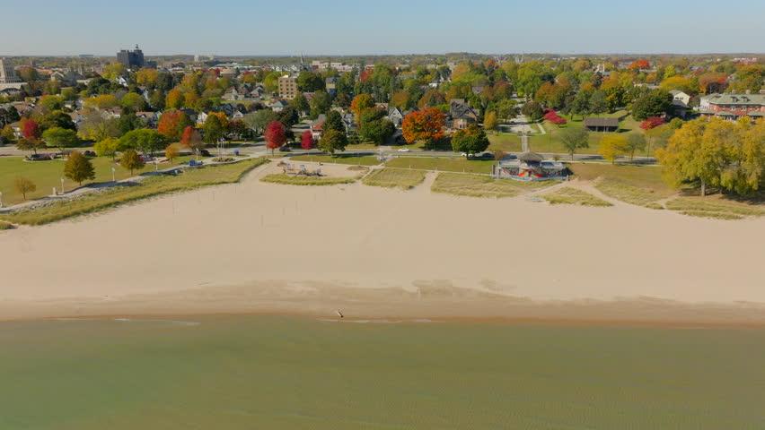 Drone aerial trucking right along the Lake Michigan shoreline in Sheboygan, Wisconsin, with neighborhood houses, colorful autumn trees, and a clear blue sky.