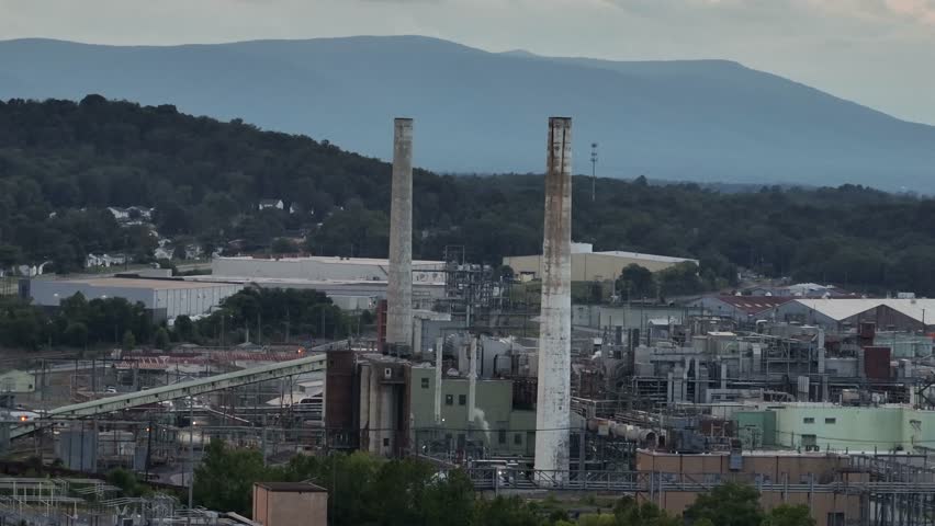 Aerial zoom of two smokestacks on industrial factory power plant in waynesboro. Virginia. Dark cloudy day in suburbia. Blue ridges mountains in background. Pipelines connecting storage.