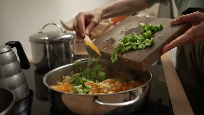 Professional chef adds chopped green bell peppers from a wooden cutting board to a pan with chicken, rice, and other vegetables while cooking on a stovetop