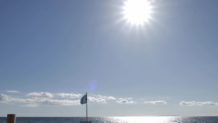Wide shot of a beautiful beach with white umbrellas, with a blue EU flag and a lifeguard station in the background on a sunny day with sun flares