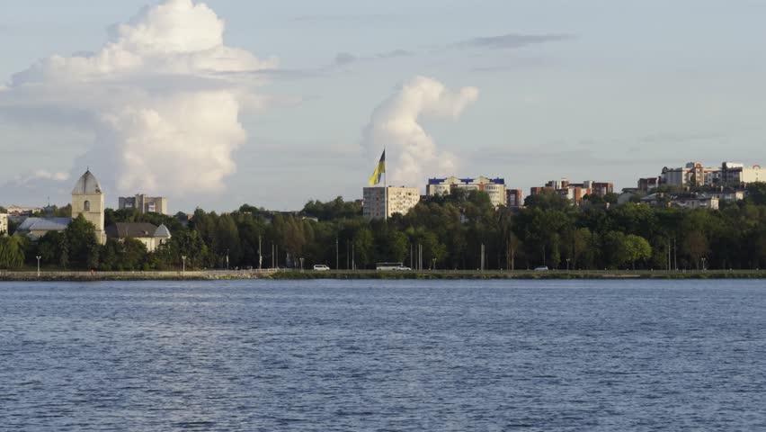 Scenic cityscape view across a wide river with buildings, trees, and a large flagpole under a sky filled with dramatic clouds.