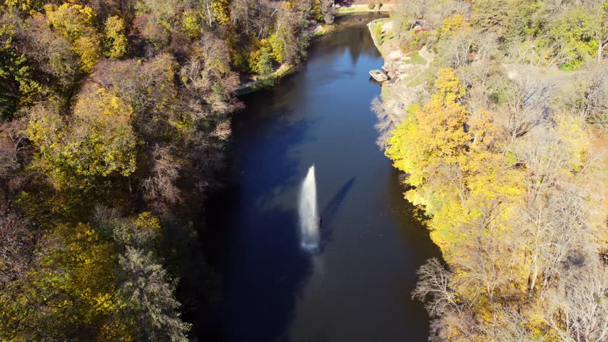 View of park on sunny autumn day. View of lake with fountain in center and trees with yellow leaves on banks in public park on sunny autumn day. Aerial drone view. Top view. Nature. Natural background