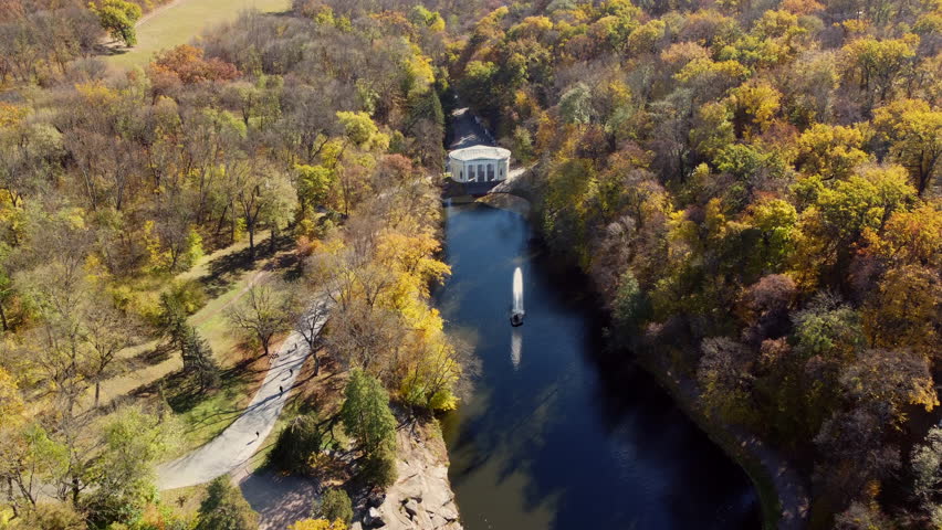 Many trees with yellow leaves, lake with fountain in center, architecture, big stones, paths walkways, people walking on paths on sunny autumn day. Aerial drone view. Panoramic autumn landscape park