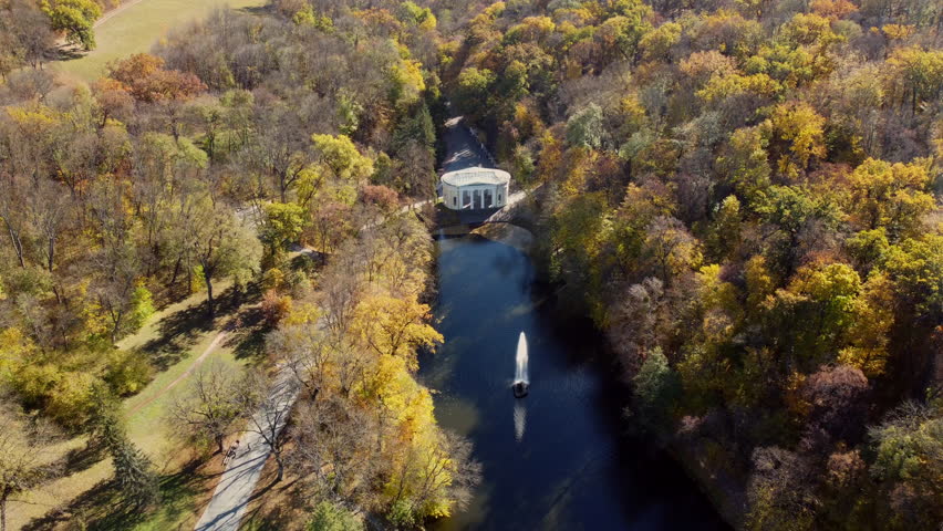 Many trees with yellow leaves, lake with fountain in center, architecture, big stones, paths walkways, people walking on paths on sunny autumn day. Aerial drone view. Panoramic autumn landscape park