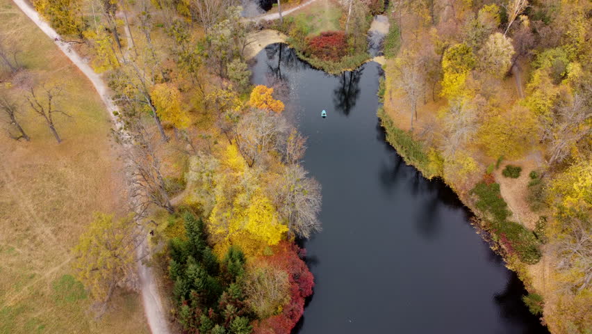 Scenery view of autumn park with trees with yellow fallen leaves, lakes, architecture, glades and people walking along dirt paths on an autumn day. Flying over autumn park. Top view. Aerial drone view