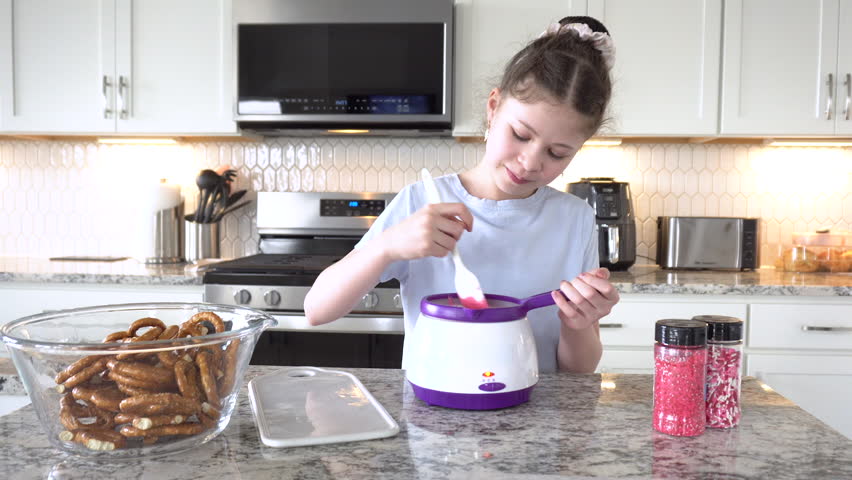With focused attention, this budding culinary artist dips pretzels into a pot of melted chocolate, creating sweet delights in the warmth of a well-lit home kitchen.