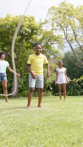Diverse family playing jump rope in sunny backyard garden on lush lawn, with shrubs. Outdoor recreation, family bonding, activity, nature, happiness, playfulness, leisure