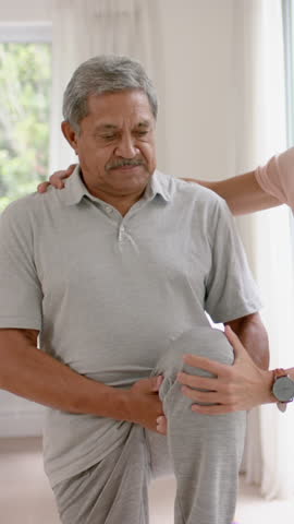 Vertical video: Senior man lifting right knee on home bench with caregiver guiding for therapy. Elderly, rehabilitation, wellness, support, mobility, care, vertical video