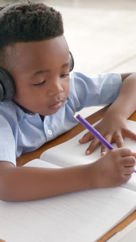 Vertical video: Boy in headphones writing in notebook at desk pausing to listen resuming writing. Youth, concentration, education, creativity, communication, modern, vertical video