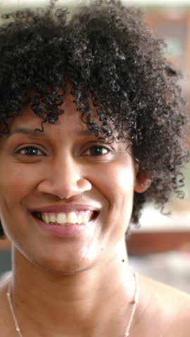 Vertical video: Woman looking at viewer, smiling then shifting into laughter at home with necklace. Joy, authenticity, expression, warmth, candid, lifestyle, vertical video