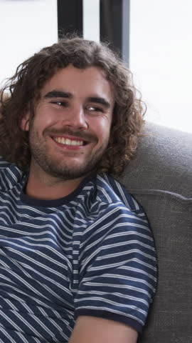 Vertical video: Hearing off-camera voice, man in t-shirt smiling, gazing on couch by window at home. Casual, relaxed, warmth, comfort, natural light, candid, vertical video