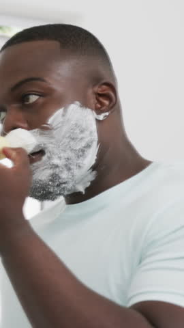 Vertical video: Morning light driving Black man applying shaving foam with brush at bathroom mirror. Masculinity, grooming, skincare, hygiene, self-care, reflection, vertical video
