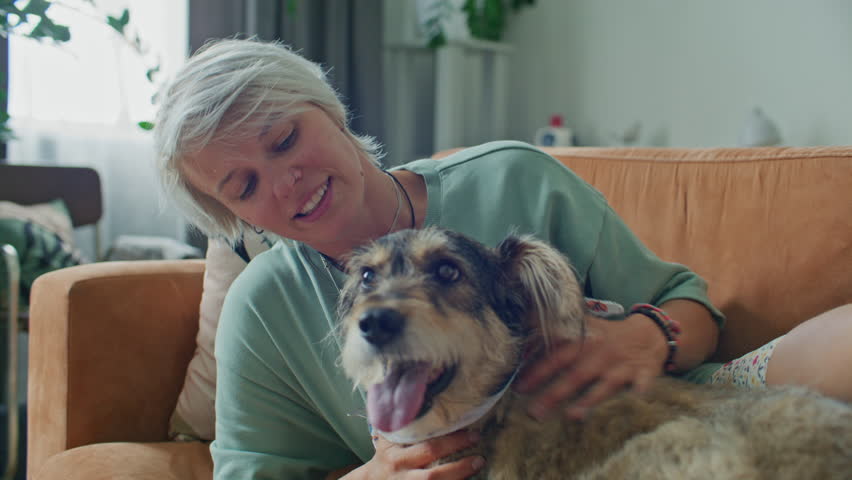 Young affectionate woman smiling while cuddling happy mixed-breed dog lying on couch in living room at home