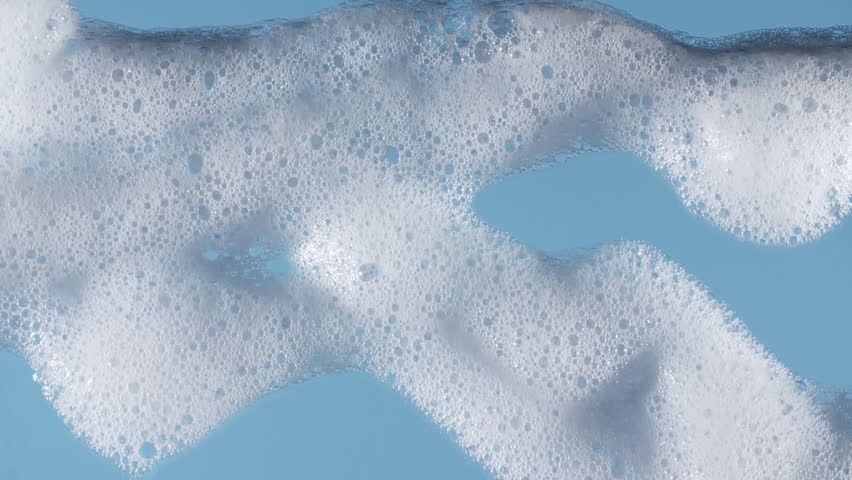 Bubbles of white soap foam. Moving Soap foam popping bubble on a blue backdrop. Soap sud macro structure. Clean, cleaning concept, laundry, shower. 