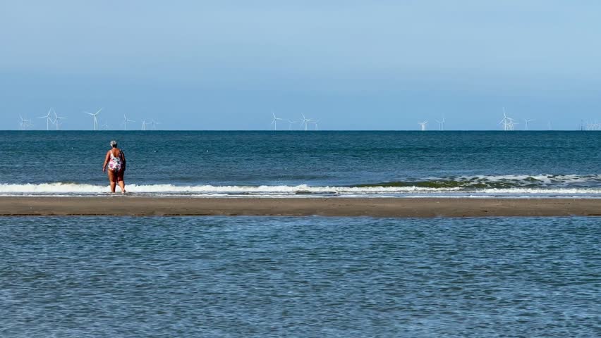 An older woman in a swimsuit walks on the beach of Noordwijk aan Zee in the ocean water outdoor at daytime during summer in The Netherlands with the windmills in the background.