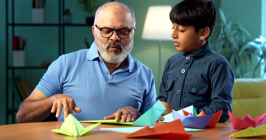 Indian grandfather and kid doing paper craft together at home, teaching colorful boat or aeroplane using bright papers, elderly man teaching grandson creative activity, family bonding indoors
