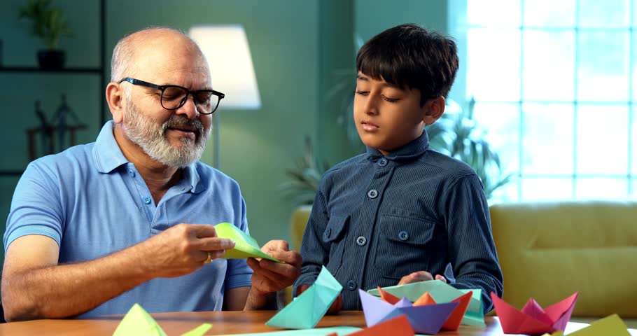 Indian grandfather and kid doing paper craft together at home, teaching colorful boat or aeroplane using bright papers, elderly man teaching grandson creative activity, family bonding indoors