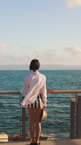 vertical shot. Slender woman in a straw hat looks at the sea and enjoys the moment. Freedom and joy. Summer vacation by the sea. Beach lifestyle.