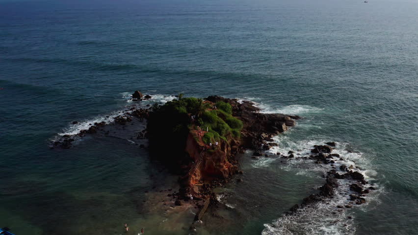 Aerial drone orbit shot circling around Parrot Rock Island in Mirissa, Sri Lanka, capturing the rocky shoreline, waves, lush greenery, and visitors on the viewpoint.