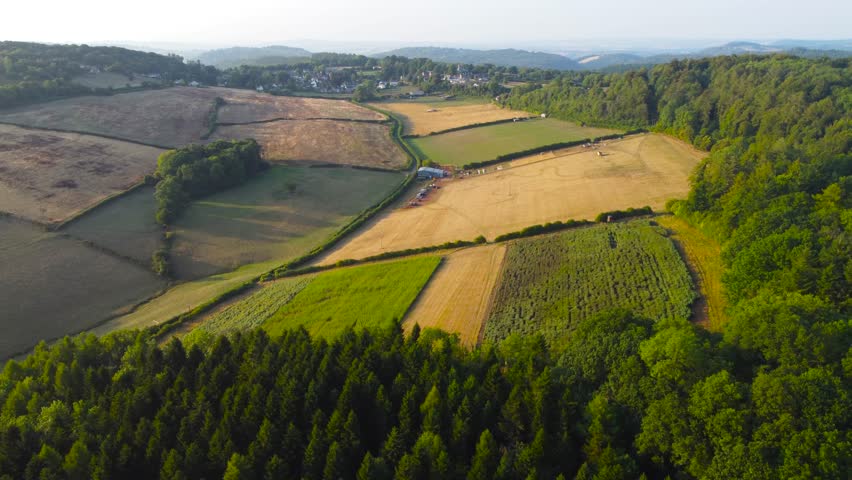 Turning Aerial View of Quaint Rural English Village Surrounded by Trees and Ploughed Farmed Fields in British Countryside. Rolling Hazy Hills in Background.