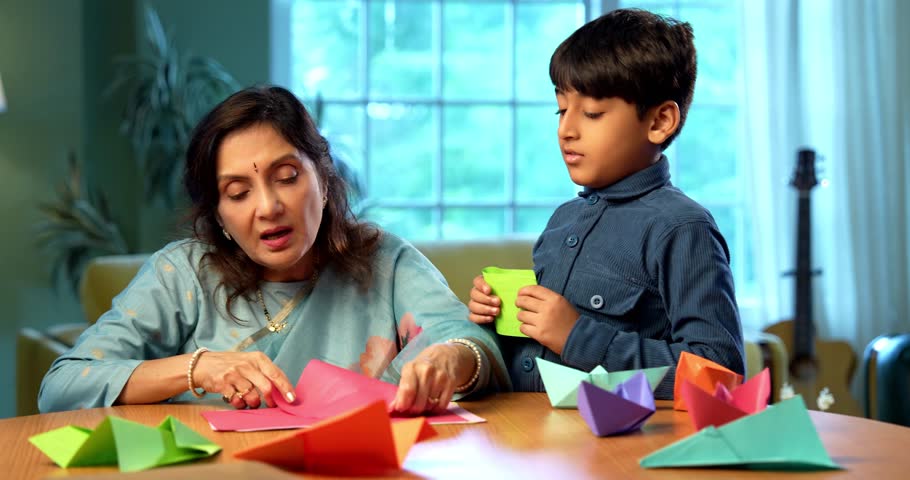 Indian grandmother and kid doing paper craft together at home, teaching colorful boat or aeroplane using bright papers, elderly man teaching grandson creative activity, family bonding indoors