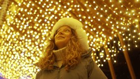 Cheerful young woman in winter attire and white furry hat gazing upward, enchanted by golden christmas lights illuminating nighttime urban street with festive glow - Powered by Shutterstock - Get 15% off with code: PIKWIZARD15