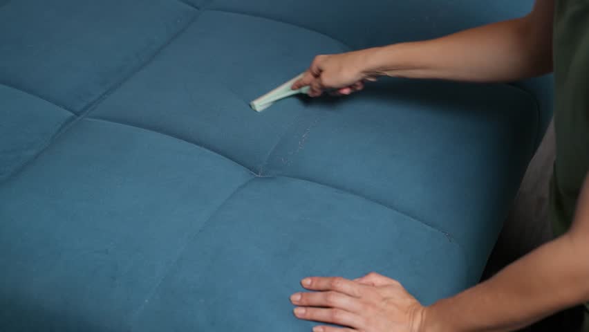 Close-up of female hand using an upholstery brush on a blue couch, scraping off dust and pet fur from the fabric. Home cleaning, sofa maintenance, housekeeping concept.