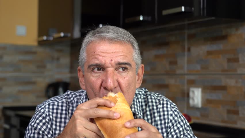 A Middle-aged Man with Short Gray Hair in a Plaid Shirt is Eating a Hot Dog at Home. Close-up of a 55-year-old man sitting at a dining table, holding a hot dog, taking a bite and chewing.
